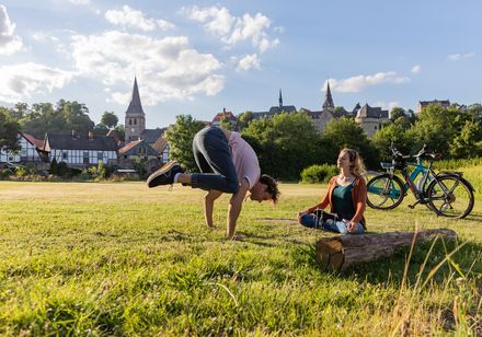 Ein Mann macht Yoga auf einer Wiese, während eine Frau mit Fahrrädern im Hintergrund sitzt.