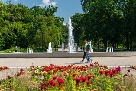 Zwei Personen spazieren an einem blühenden Blumenbeet vor einem großen Springbrunnen im Park vorbei.