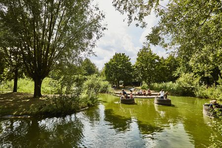 Menschen fahren mit Rundbooten auf grünlichem Wasser durch einen idyllischen, von Bäumen gesäumten Kanal.