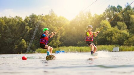 Zwei Kinder auf Wakeboards, gezogen von einem Seil über die Wasseroberfläche, unter strahlendem Sonnenschein.