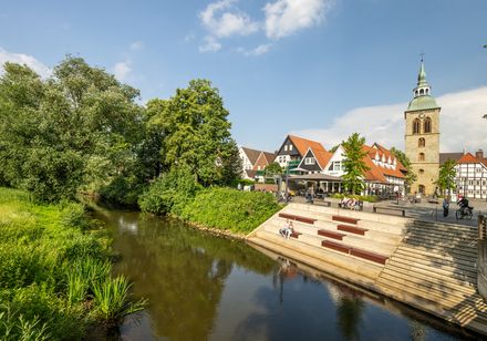 Historische Fachwerkhäuser und eine Kirche am Flussufer mit grüner Vegetation und klarer Himmel.