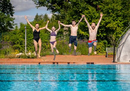 Vier Personen springen fröhlich Hand in Hand in ein Freibadbecken an einem sonnigen Tag.