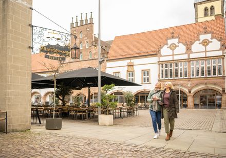 Zwei Frauen spazieren durch einen historischen Marktplatz mit Fachwerkhäusern und einem Café.
