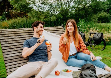 Ein junges Paar sitzt auf einer Bank im Grünen, isst Obst und genießt einen entspannten Moment.