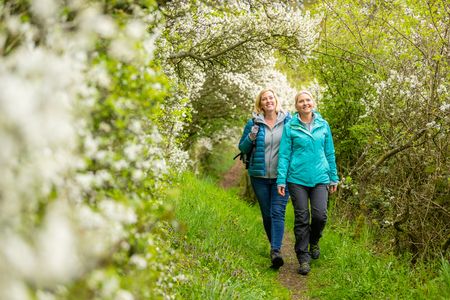 Zwei Frauen wandern lächelnd auf einem blühenden, grünen Pfad, umgeben von blühenden Sträuchern.
