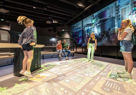 3 Kinder bei Erlebnisstation, ein Kind springt, Eltern im Hintergrund auf einer Bank