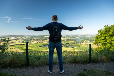Ein Mann steht auf einem Hügel mit ausgestreckten Armen und blickt auf eine weite Landschaft.