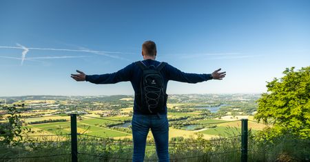 Ein Mann steht auf einem Hügel mit ausgestreckten Armen und blickt auf eine weite Landschaft.