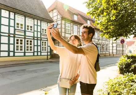 Junges Paar macht Selfies vor historischen Fachwerkhäusern in einer sonnigen, baumgesäumten Straße.
