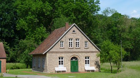 Historisches Backsteinhaus mit rotem Satteldach, umgeben von grünen Bäumen und Wiesen in ländlicher Umgebung.