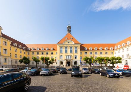 Stadtschloss Cottbus im Barockstil mit rotem Dach, umgeben von parkenden Autos auf dem Vorplatz.