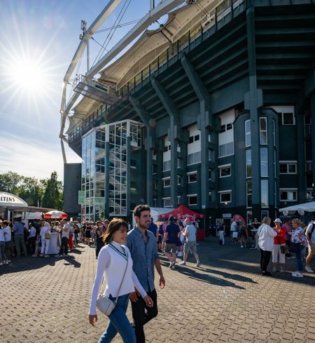 Menschen spazieren bei Sonnenschein vor einem großen Stadion, umgeben von Besuchern und Verkaufsständen.