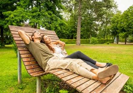 Ein Paar entspannt sich auf einer Holzbank in einem grünen Park unter sonnigem Himmel.