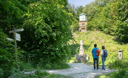 Zwei Wanderer stehen auf einem Waldweg und blicken auf einen steinernen Gedenkstein.