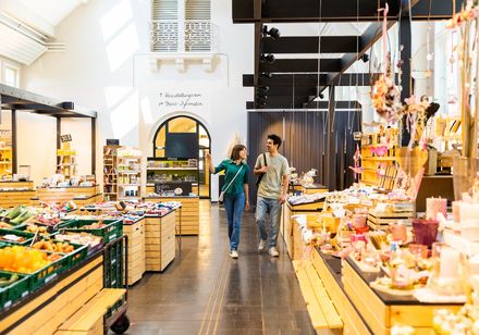 Ein Paar spaziert durch einen modernen Markt mit frischem Obst, Blumen und verschiedenen Produkten.