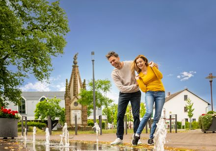 Ein glückliches Paar spielt im sonnigen Stadtpark von Rahden mit sprudelnden Wasserfontänen.