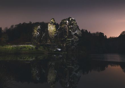 Ein stimmungsvoller Abendblick auf die beleuchteten Externsteine, die sich im Wasser spiegeln.