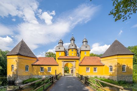 Gelbes Barockschloss mit Türmen, Backsteinweg führt zur Holzbrücke, Bäume und blauer Himmel im Hintergrund.