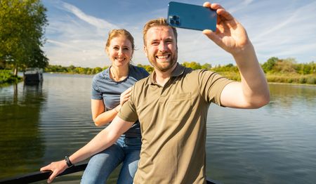 Ein lächelndes Paar steht auf einem Steg und macht ein Selfie vor einem See mit blauem Himmel.