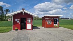Zwei rote Holzhütten in ländlicher Umgebung, eine beschriftet mit "SOFTEIS", flankiert von Feldern.