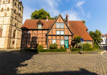 Fachwerkhaus mit roten Ziegeln, markanten Holzbalken und Satteldach, neben einer historischen Kirche.