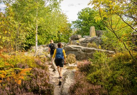 Zwei Wanderer auf einem steinigen Pfad, umgeben von üppiger Vegetation und einem Obelisken im Hintergrund.