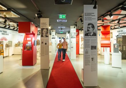 Ausstellungshalle mit roten Teppich, historischen Spielautomaten und Informationstafeln an weißen Säulen.