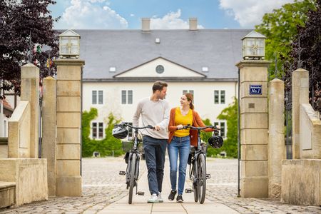 Ein Paar schiebt Fahrräder durch ein elegantes Tor vor einem historischen Gebäude bei Sonnenschein.