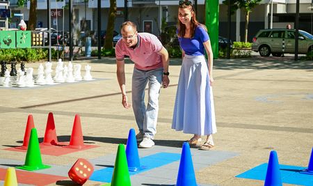 Menschen spielen ein übergroßes "Mensch ärgere dich nicht"-Spiel im Freien auf einem Platz.