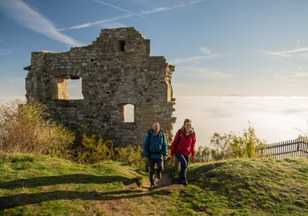 Zwei Wanderer erklimmen einen Hügel mit einer alten Steinruine im Hintergrund, umgeben von Nebel.