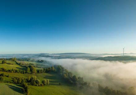Panorama von Nebelfeldern über grüner Hügellandschaft, mit Windrädern am rechten Bildrand.
