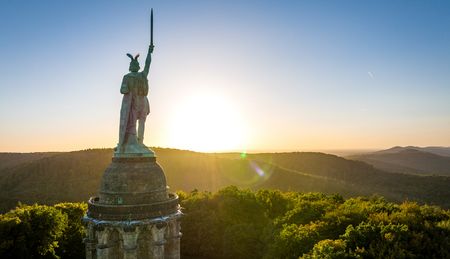 Sonnenuntergang hinter dem Hermannsdenkmal im Teutoburger Wald, umgeben von bewaldeten Hügeln.