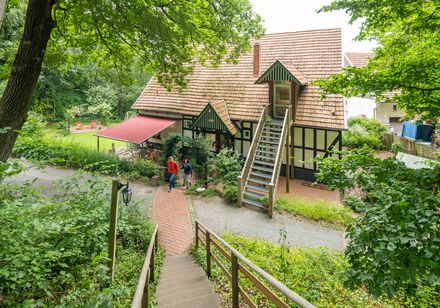 Fachwerkhaus im Grünen mit rotem Vordach, umgeben von Bäumen und Spaziergängern auf Weg.