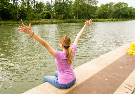 Frau im rosa T-Shirt sitzt glücklich am Flussufer, breitet die Arme aus, umgeben von grüner Natur.