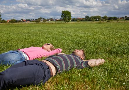 Zwei Personen liegen entspannt auf einer grünen Wiese unter einem teils bewölkten Himmel.