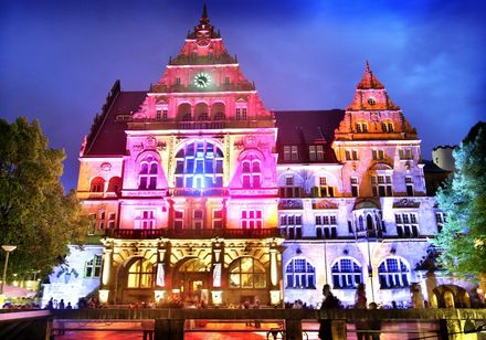 Historisches Gebäude mit neugotischer Fassade, nachts bunt beleuchtet, vor einem klaren Himmel.