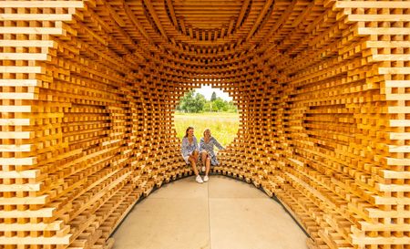 Zwei Frauen sitzen in einem Holzkubus aus gekreuzt geschichteten Holzlatten