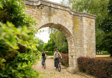 Zwei Radfahrer durchqueren einen historischen steinernen Torbogen in einer grünen Parklandschaft.