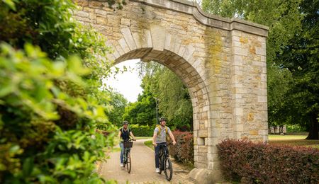 Zwei Radfahrer durchqueren einen historischen steinernen Torbogen in einer grünen Parklandschaft.