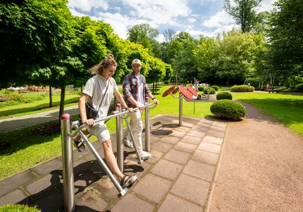 Zwei Personen nutzen Fitnessgeräte im Freien, umgeben von grünen Bäumen und einem gepflegten Weg im Kurpark.