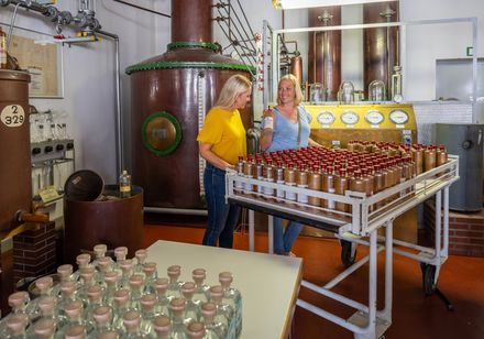 Zwei Frauen besichtigen eine Destillerie mit Glasflaschen und kupfernen Brennblasen im Hintergrund.
