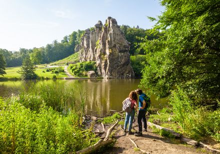 Zwei Wanderer betrachten die imposanten Externsteine am Ufer eines Sees inmitten grüner Natur.