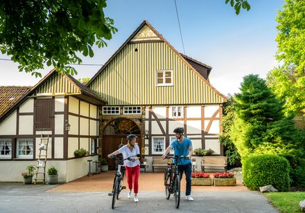 Zwei Menschen schieben Fahrräder vor einem traditionellen Fachwerkhaus in grüner Umgebung entlang.