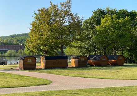 Sechs holzverkleidete Campingfässer stehen auf einer Wiese neben einem See, umgeben von Bäumen.