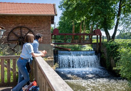 Ein Paar betrachtet von einer Holzbrücke ein kleines Wasserkraftwerk mit plätscherndem Wasserfall.