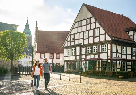 Fachwerkhäuser auf gepflastertem Platz bei Sonnenschein, Paar spaziert vorbei.