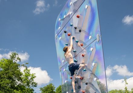 Ein Mann klettert an einer transparenten Kletterwand über einem Außenpool, umgeben von Bäumen.