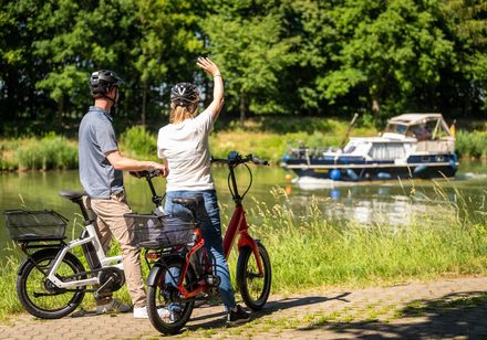 Zwei Radfahrer am Flussufer, winken einem vorbeifahrenden Boot, umgeben von grüner Natur.