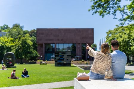 <p>Paar sitzt auf einer Mauer, schaut auf modernen Museumsbau in grünem Park, bei Sonnenschein.</p>
