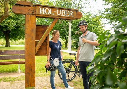 Ein Mann und eine Frau stehen mit Fahrrädern vor einem Hol-Über-Schild im Grünen und lächeln.